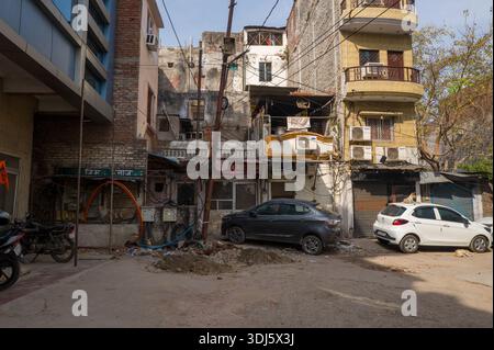 Lucknow, India - March 16, 2024: The image shows the facades of old and modern buildings along a narrow lane with parked cars, motorcycles, visible shopfronts with Hindi signage, exposed electrical wiring, air conditioning units, and debris on the ground under daylight. Stock Photo
