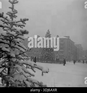 Sloviansk, Donetsk region, Ukraine, USSR. 1970s. Winter holiday scene on the Square of the October Revolution (now Soborna Square). A large decorated New Year tree stands in the center, surrounded by a wooden fence. Citizens walk around the snowy square. In the foreground, a blurry snow-covered pine branch frames the view, adding depth to the composition. In the background, a residential building with a shop on the ground floor is visible. The image captures the festive atmosphere and urban life of the peaceful Donbas. An archival black and white photograph. Stock Photo