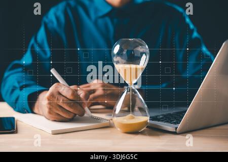 Man writing ideas in book, watching hourglass time for business work, sandglass showing hour countdown, focused on laptop for planning, managing deadl Stock Photo