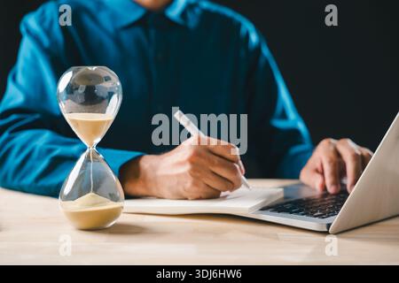 Hourglass shows passing time, business man focused on work, diligently writing notes in book and using laptop, managing hour glass sand countdown and Stock Photo