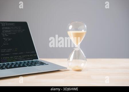 Deadline approaching for critical business task, hourglass glass timer shows sand flowing next to laptop computer displaying code, symbolizing importa Stock Photo