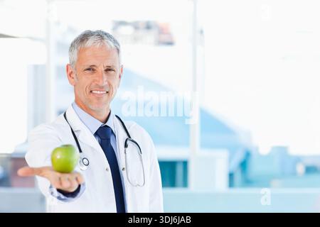 Mature male doctor holding green apple toward camera at clinic with lab coat and stethoscope. Physician, medical, clinician, portrait, backlit, bright Stock Photo