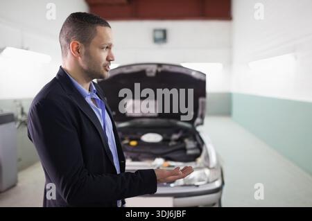 Adult man wearing blazer, blue shirt gesturing beside car with raised hood in garage, copy space. Workshop, bay, automotive, engine, diagnostic, indus Stock Photo