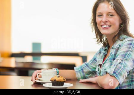 Woman in her 20s wearing plaid shirt smiling at camera holding cup and muffin, copy space. Cafe, backlit, pastry, beverage, casual, candid, warm Stock Photo