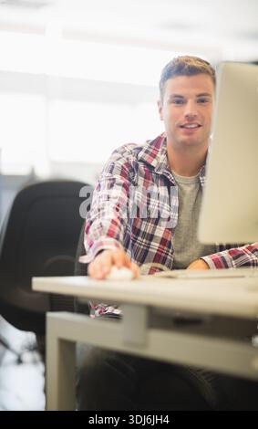 Twenty-something man wearing red plaid shirt sitting at desk in bright lab using mouse and monitor. Office, workspace, screen, keyboard, ergonomic, ba Stock Photo
