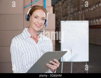 Mid-adult female wearing headset and striped shirt, holding clipboard and pen in warehouse. Inventory, logistics, supervisor, coordinator, storage, fa Stock Photo