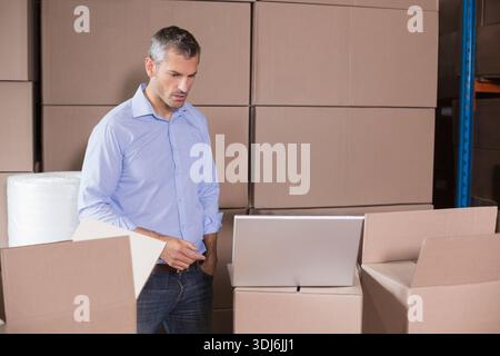 Mid-adult man inspecting silver laptop in warehouse with boxes and bubble wrap, in light blue shirt. Storage, logistics, packaging, inventory, cartons Stock Photo