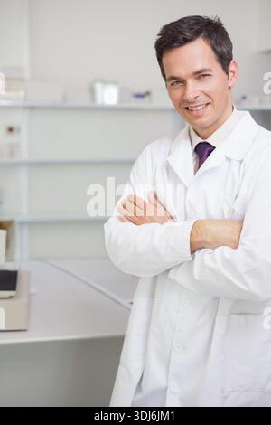 Mature man in white lab coat standing arms crossed beside benchtop analyzer and shelves, copy space. Professional, scientist, clinical, laboratory, mi Stock Photo