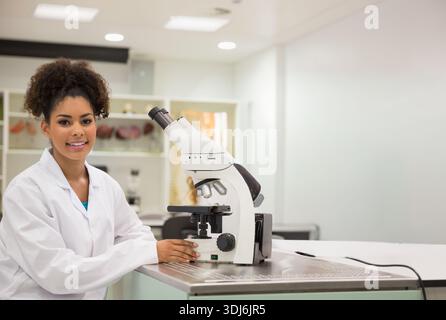 African American woman wearing lab coat working at compound microscope using focus knob, copy space. Scientist, laboratory, workbench, stainless, cabi Stock Photo