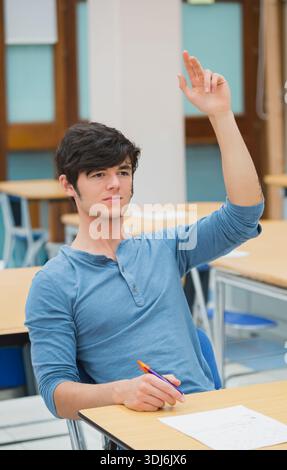 Male student in blue henley raising right hand holding purple pen orange cap papers at desk. Learner, classroom, participation, engagement, curiosity, Stock Photo