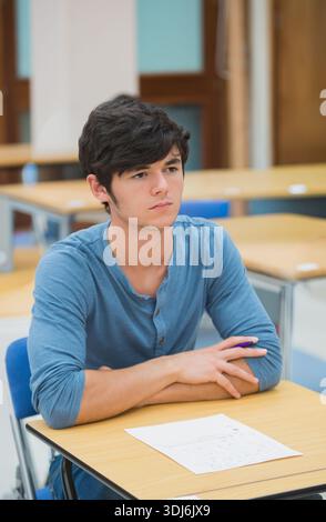 Male student sitting at class desk wearing blue henley holding purple pen reading handwritten notes. Learner, classroom, focus, concentration, study, Stock Photo