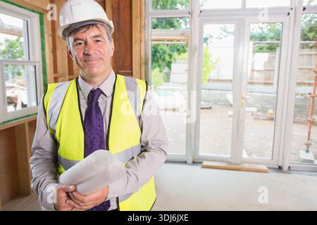 Senior man wearing white hard hat and hi-vis vest holding rolled blueprints inside renovated room. Construction, renovation, scaffolding, drawings, su Stock Photo