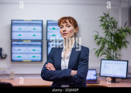 Professional woman standing in control room, wearing navy blazer, monitoring multi-panel dashboards. Executive, analyst, data, screens, office, manage Stock Photo