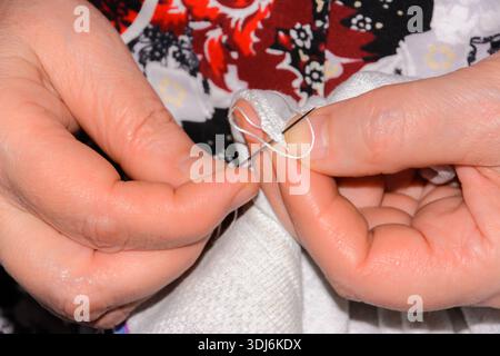 An elderly woman sews things up using threads and a needle. Clothes torn and a hole formed. Clothing repair. The hands of an old woman stitching polo. Stock Photo