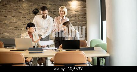 A group of four professionals collaborates around a table in a contemporary office setting. They are sharing documents and ideas, clearly engaged in a Stock Photo