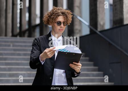 Happy Caucasian businessman work with documents reading information business papers prepare financial report. Young guy man in suit analyzing graph documents, calculate taxes standing outside office. Stock Photo