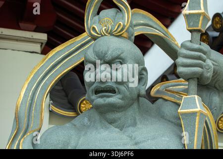 A close-up of a fierce stone statue depicting a guardian figure with an intense expression, ornate details, and a staff, set against a traditional arc Stock Photo