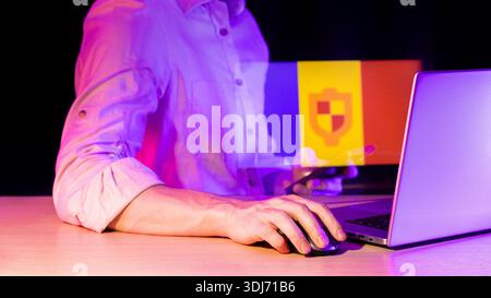 Close-up of hand on mouse near laptop, dark background with purple lighting, holographic display of colorful vertical striped flag and security shield Stock Photo