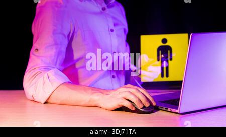 Close-up of hand operating mouse beside laptop, holographic warning icon of male figure with device in dim purple glow. Stock Photo