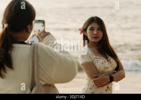 Khao Lak, Thailand, 16 11 2025: real authentic moments of life- Woman taking a picture her Thai friend on her smartphone against the sunset at sea Stock Photo