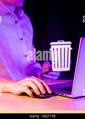 Close-up of hand holding mouse near laptop displaying glowing trash can symbol in purple neon atmosphere. Stock Photo