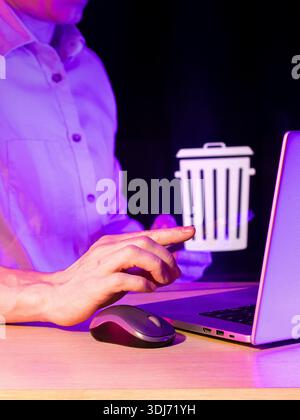 Side view of hand pinching white floating delete icon in dimly lit purple environment with laptop and computer mouse on desk. Stock Photo