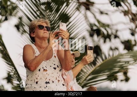 Khao Lak, Thailand, 16 11 2025: real authentic moments of life- An adult elderly woman in sunglasses photographs a beautiful sunset against a Stock Photo