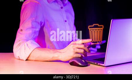 Close-up of male hand pointing at glowing orange trash symbol while working on laptop with mouse on desk in dark room with purple neon glow Stock Photo