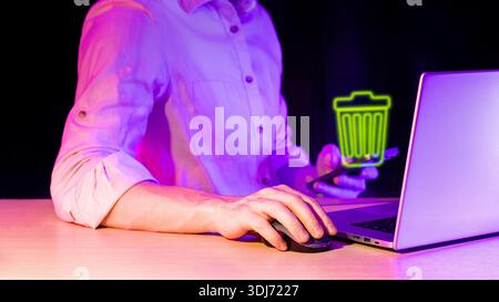 Hand operating computer mouse beside glowing green trash hologram while male works on laptop at wooden desk in purple-lit dark space Stock Photo