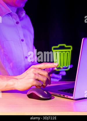 Close view of male hand with extended finger touching glowing green trash symbol near open laptop and mouse on desk in neon purple ambiance Stock Photo