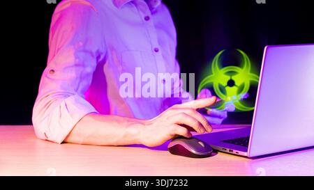 Extended male finger directing towards glowing green biohazard hologram beside open laptop and computer mouse on desk in purple-lit darkness Stock Photo