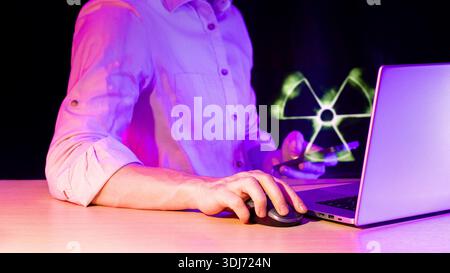 Close-up of hand using mouse on desk with laptop and phone displaying neon radiation hazard icon in dark setting Stock Photo