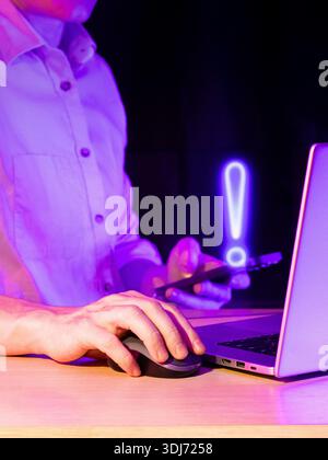 Close view of user gripping smartphone displaying neon blue alert symbol next to computer in low light Stock Photo