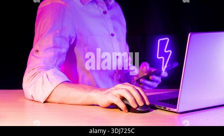 Workspace scene with person holding phone showing purple lightning symbol while operating laptop Stock Photo