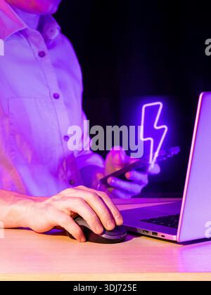 Man using mouse on desk with laptop and smartphone displaying vibrant purple bolt in shadowy room Stock Photo