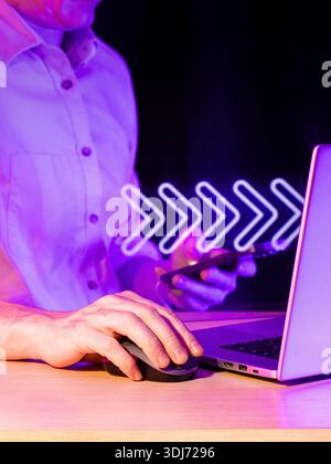 Hands engaged with mouse and mobile, featuring neon right arrows icon in futuristic digital workspace under dim conditions Stock Photo