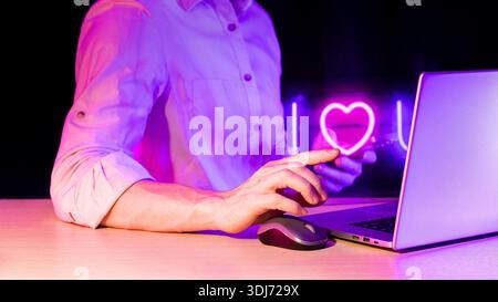 Close-up of hand holding smartphone pointing toward glowing neon I love you sign while other hand rests on computer mouse beside laptop in purple ligh Stock Photo