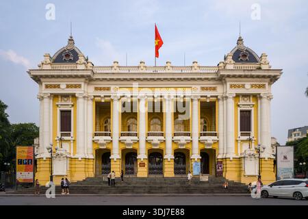 Hanoi, Vietnam - September 13, 2022: The facade of the Hanoi Opera House features yellow and white colonial architecture, a Vietnamese flag on the roof, and several people standing or walking on the front steps. Stock Photo