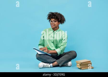 Smart black teen guy in glasses taking notes, holding notepad and pen, sitting with books nearby on blue background Stock Photo