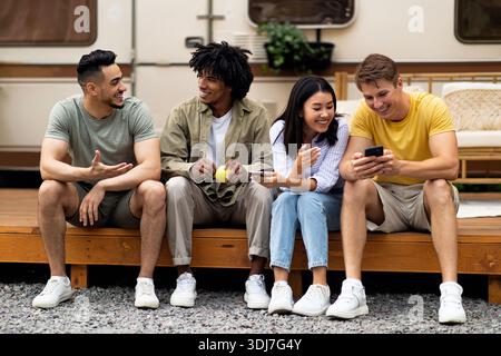 Diverse friends sitting near motorhome, talking to each other, using smartphone, spending time together on camping trip Stock Photo