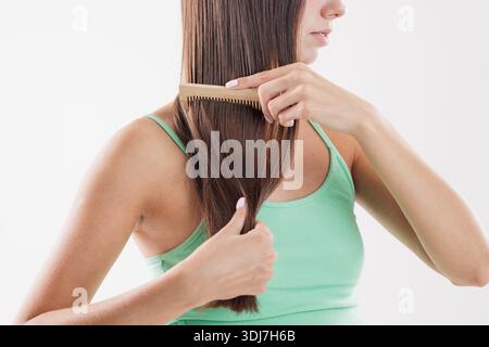 Young woman combing long straight brown hair with a wooden comb, promoting natural beauty, self-care, and healthy hair care routine on a white backgro Stock Photo