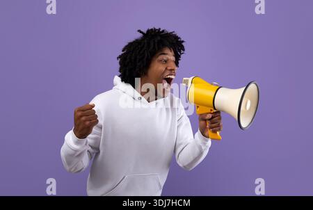 Emotional black adolescent shouting into megaphone, making announcement over violet studio background Stock Photo