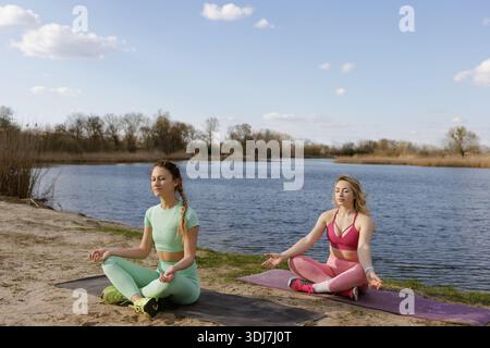 Two young women meditating outdoors near a calm river on yoga mats under a clear blue sky, practicing mindfulness. Stock Photo