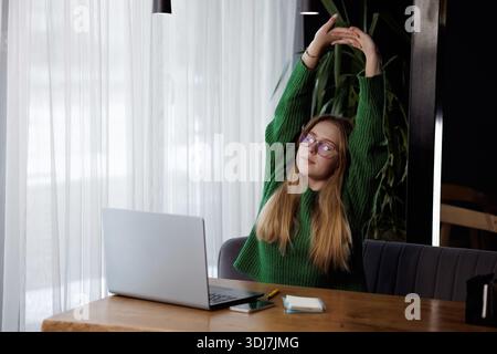 Young woman in green sweater stretching arms during remote work break at desk with laptop and coffee. Stock Photo