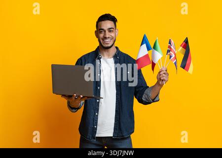 Cheerful arab guy holding laptop and international flags on yellow Stock Photo