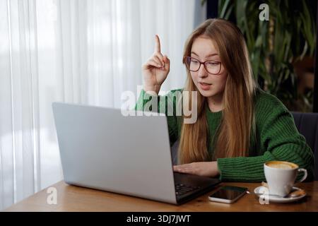 Smart young woman in glasses raising finger with idea while video conferencing on laptop in a cozy cafe. Stock Photo