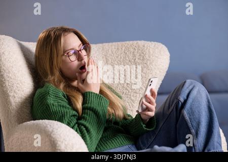 Sleepy young woman yawns while holding smartphone, feeling tired and drowsy after screen time at home. Stock Photo