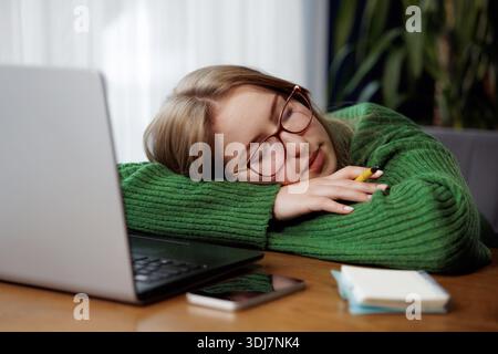 Tired young woman in glasses falling asleep at desk with laptop and notepad, exhausted while working or studying. Stock Photo