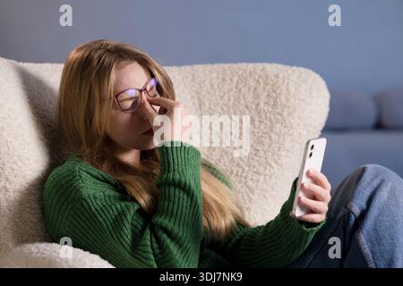 Young woman with glasses rubs eyes while using smartphone, feeling digital fatigue and eye strain at home. Stock Photo