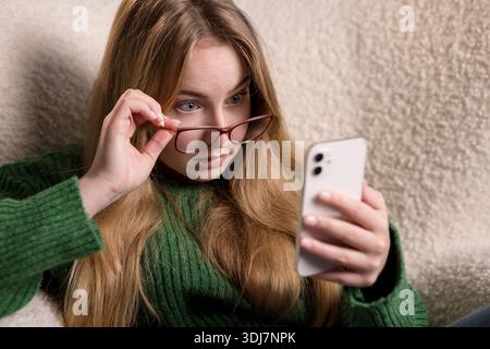 Surprised young woman lifting glasses and staring at smartphone screen, reacting to unexpected message. Stock Photo